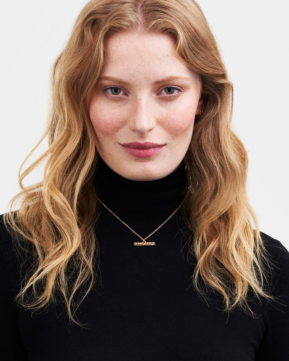 Woman with wavy blonde hair wearing a black turtleneck and gold necklace on a white background