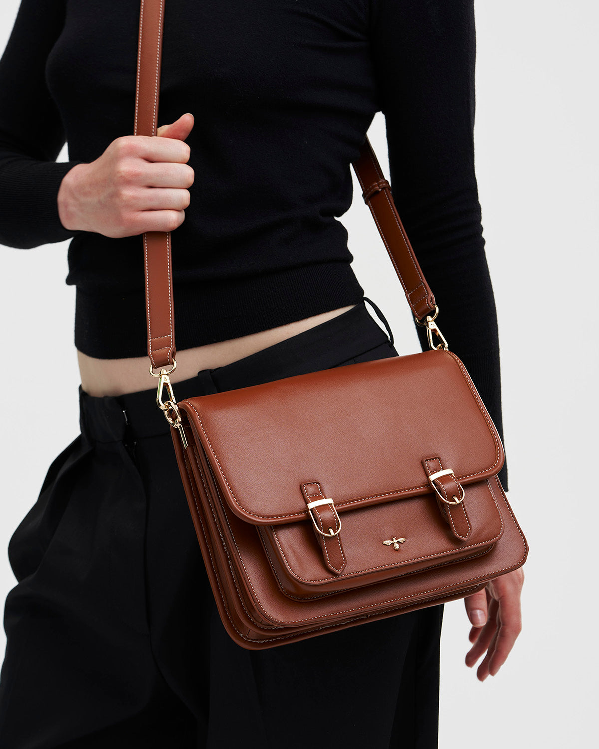 Brown leather satchel bag held by a person wearing a black outfit on a light gray background