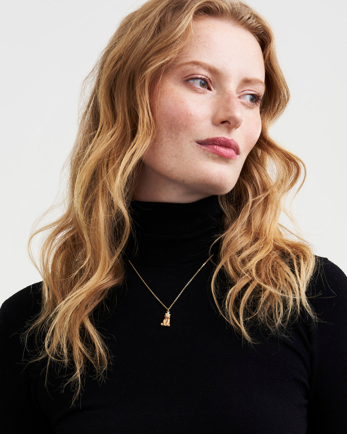 Woman wearing a black turtleneck and gold necklace with a small pendant against a white background