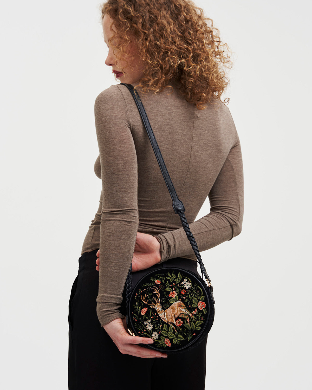 Woman holding a black round bag with floral and deer embroidery on a white background