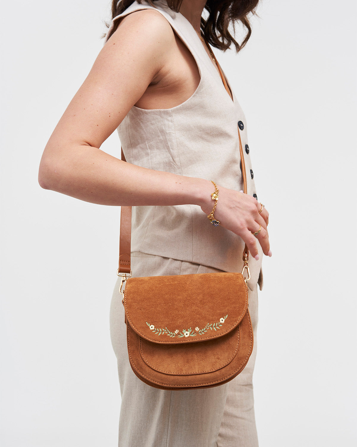 Person wearing a beige sleeveless top and holding a brown leather bag with floral embroidery on a white background