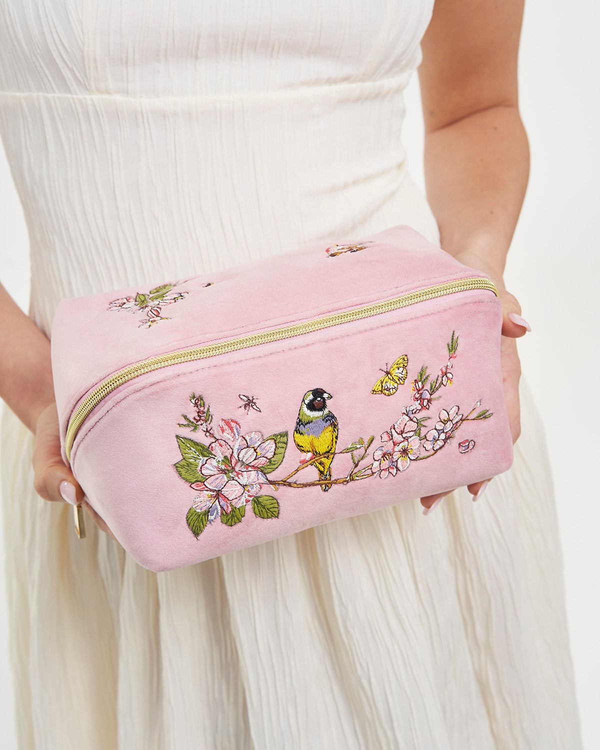 Pink pouch with floral and bird embroidery held by a person wearing a white dress.