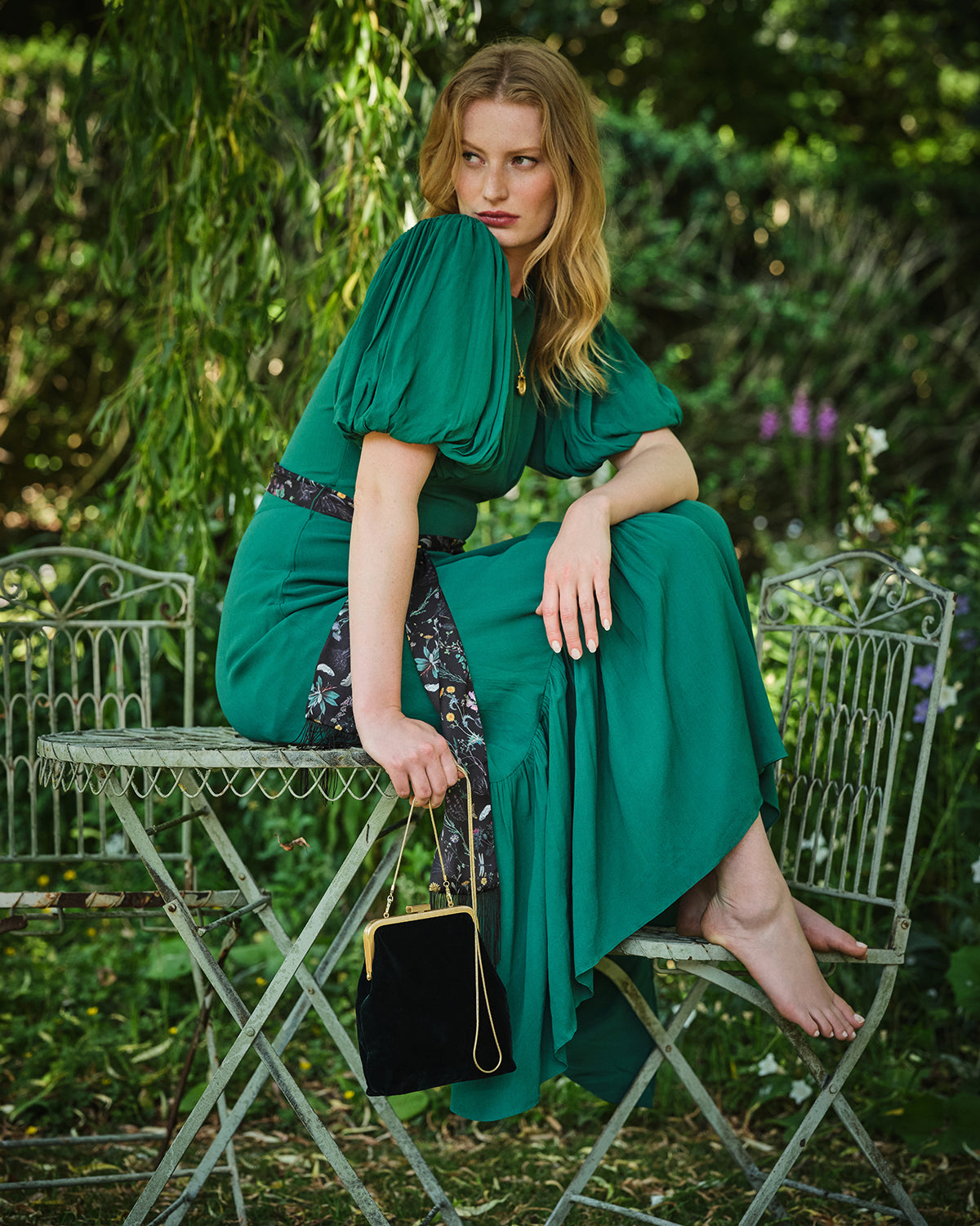 Woman in a green dress sitting on a metal chair outdoors with a blurred natural background