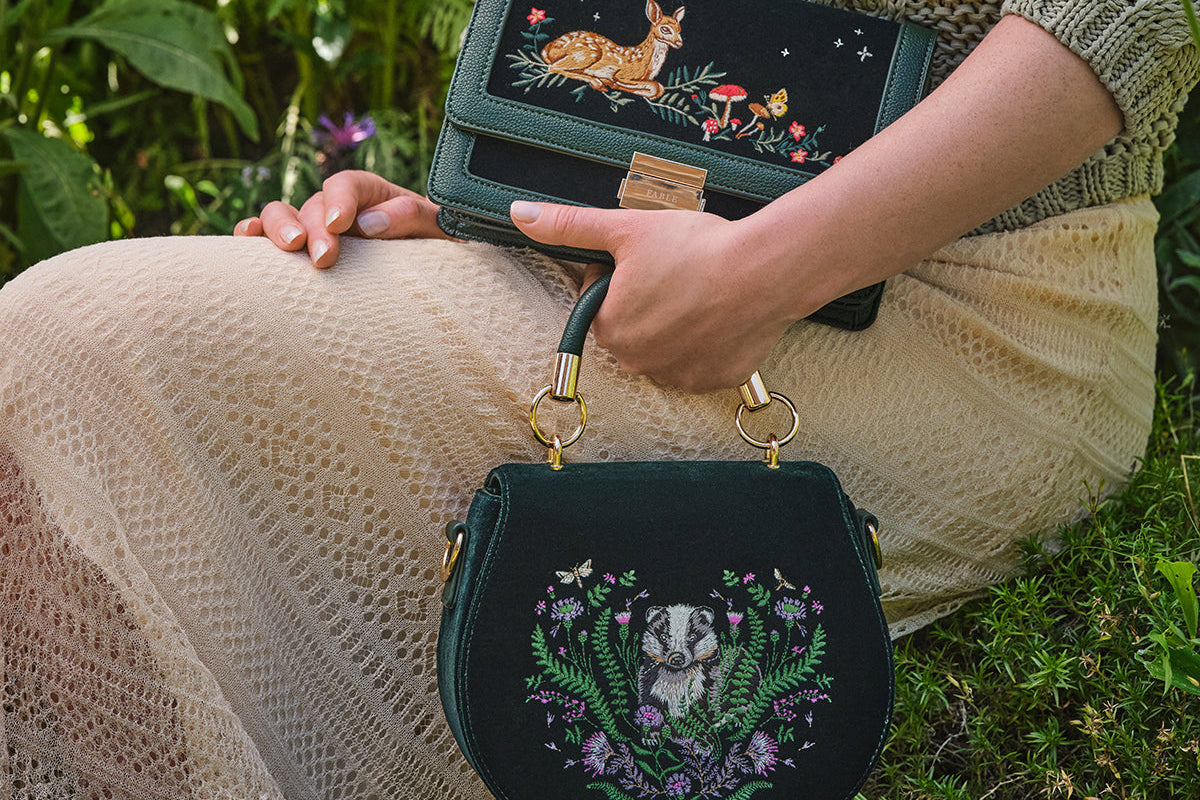Person holding a dark green handbag with embroidered designs in a natural setting