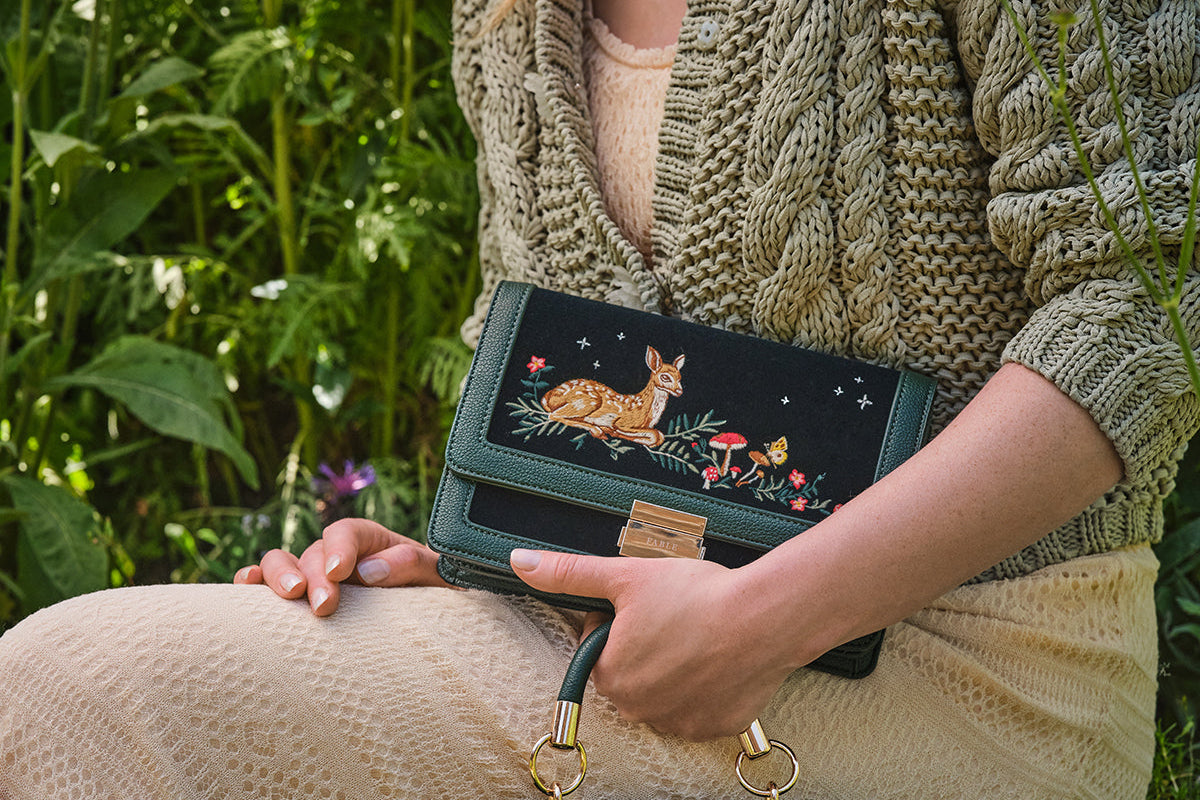 Person holding a dark green handbag with embroidered designs in a natural setting
