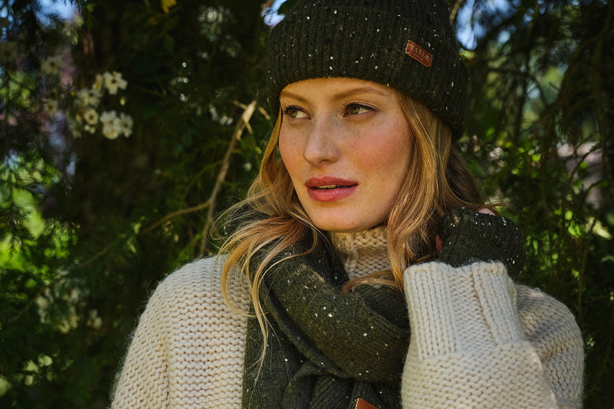 Woman wearing a beige sweater, black knit hat, and dark scarf outdoors with greenery in the background