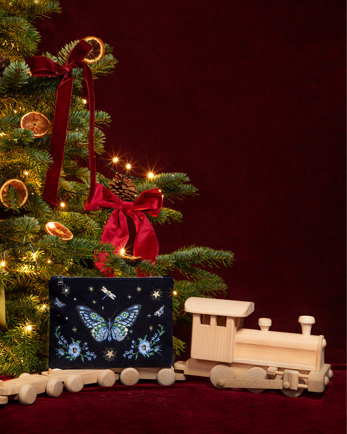 A dark Christmas-themed gift box with a blue butterfly and floral design sits on a red surface next to a wooden toy train, with a decorated Christmas tree in the background.