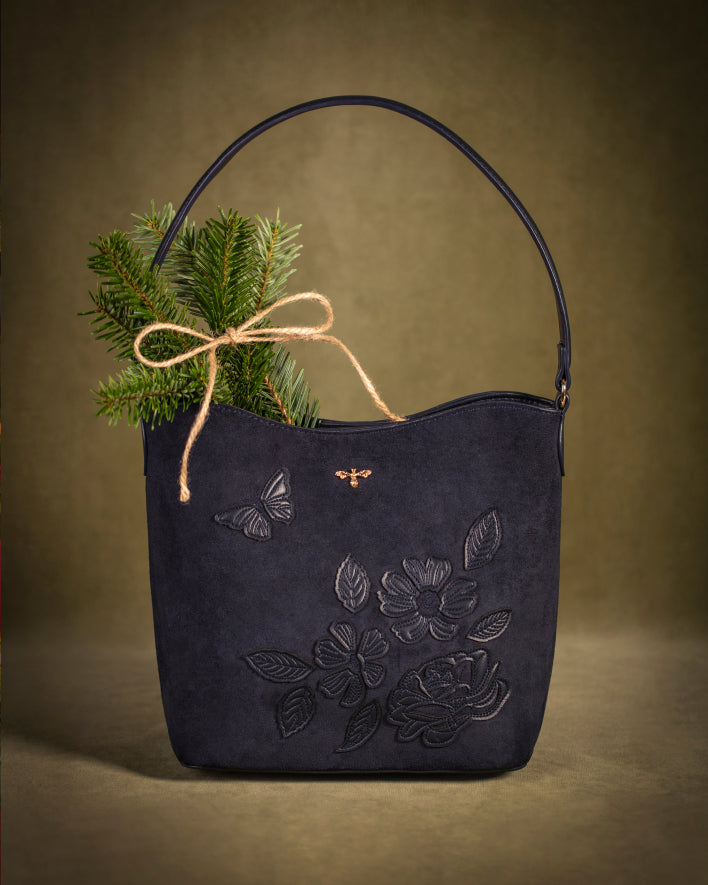 A dark navy blue suede handbag with embossed floral and butterfly details, featuring a natural jute twine bow with green pine branches tucked into the side, set against a warm brown studio background.