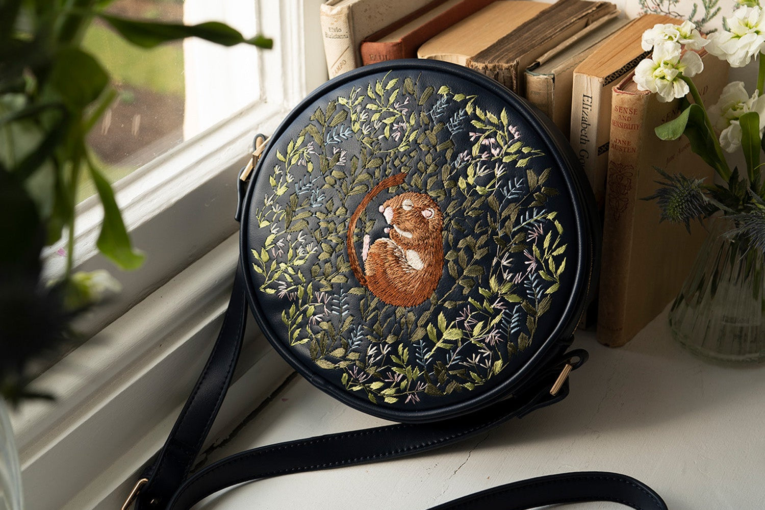 Round embroidered bag with a sleeping dormouse design on a surface near books and a plant.
