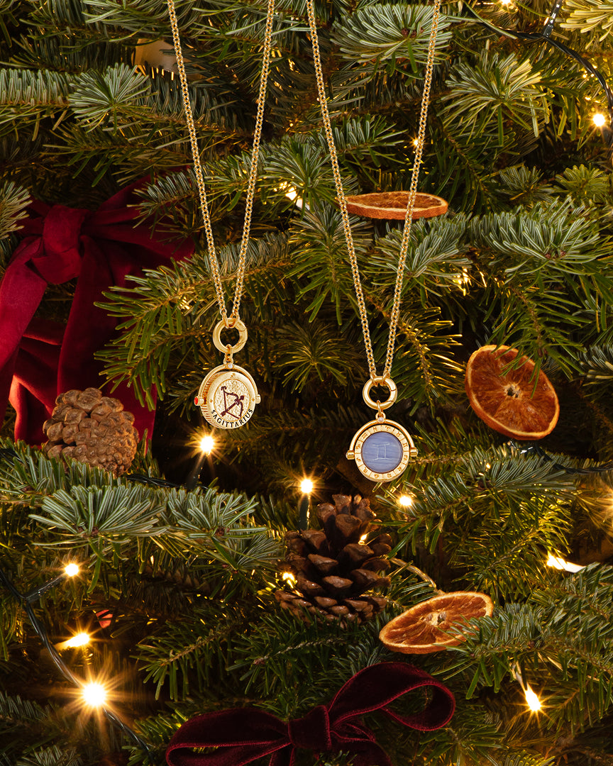 Two gold necklaces with round pendant charms hang from a Christmas tree decorated with lights, pinecones, dried orange slices, and red ribbon bows. One pendant is white with a black symbol, and the other is blue with a gold border.