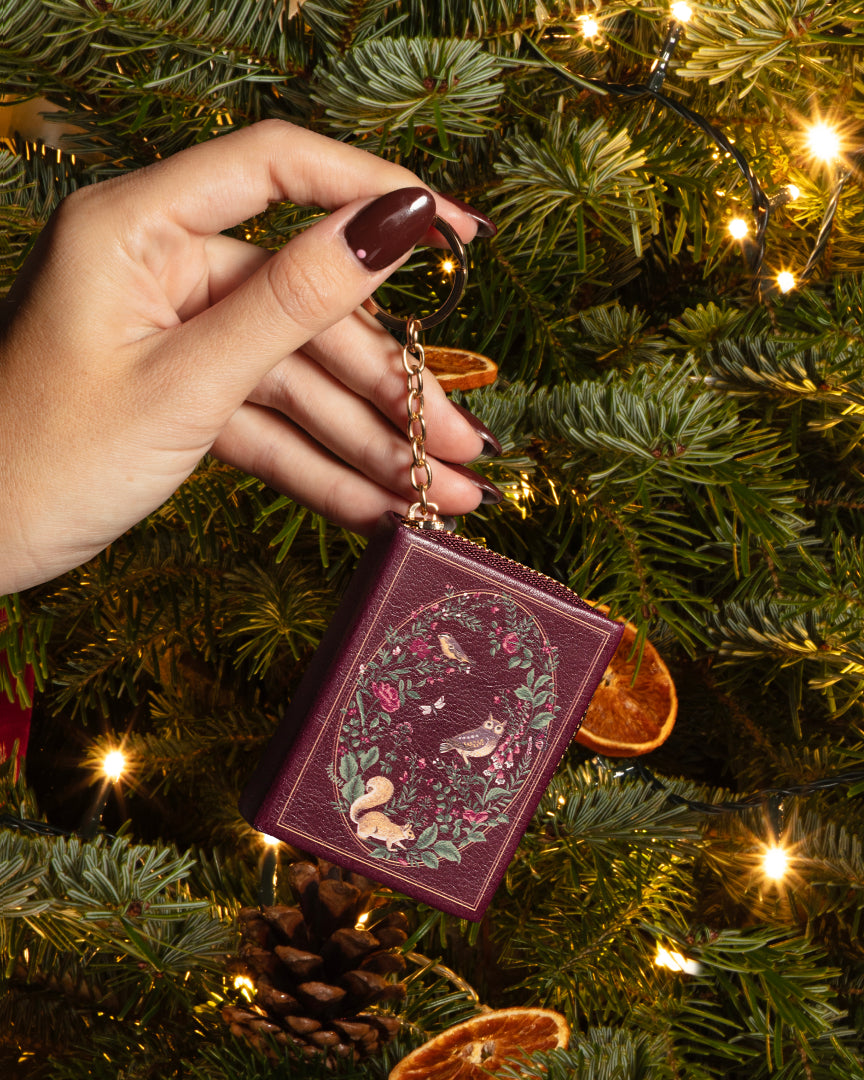 A hand with dark burgundy nail polish holds a small decorative book-shaped keychain in front of a lit Christmas tree. The keychain has a burgundy cover with an embroidered design of birds, flowers, and foliage, attached to a gold chain.