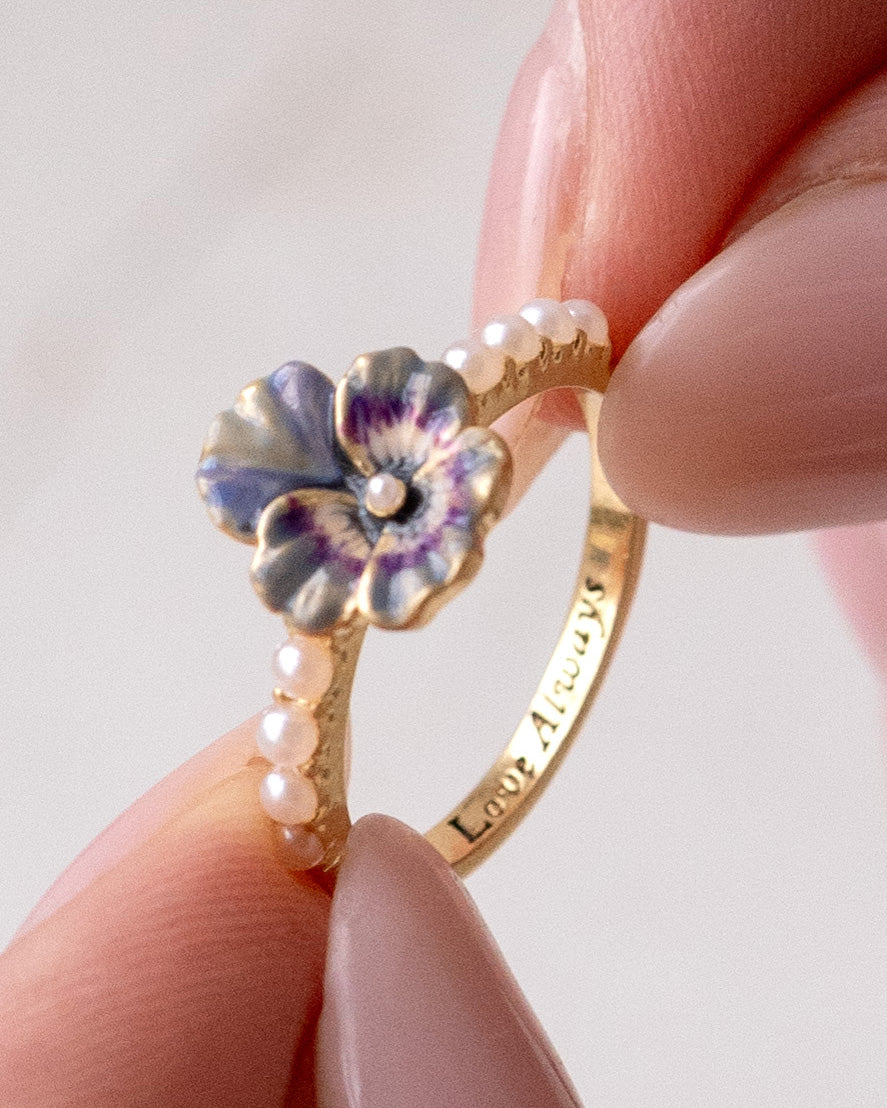 Gold ring with a floral Pansy design and pearl detail held between fingers against a light background