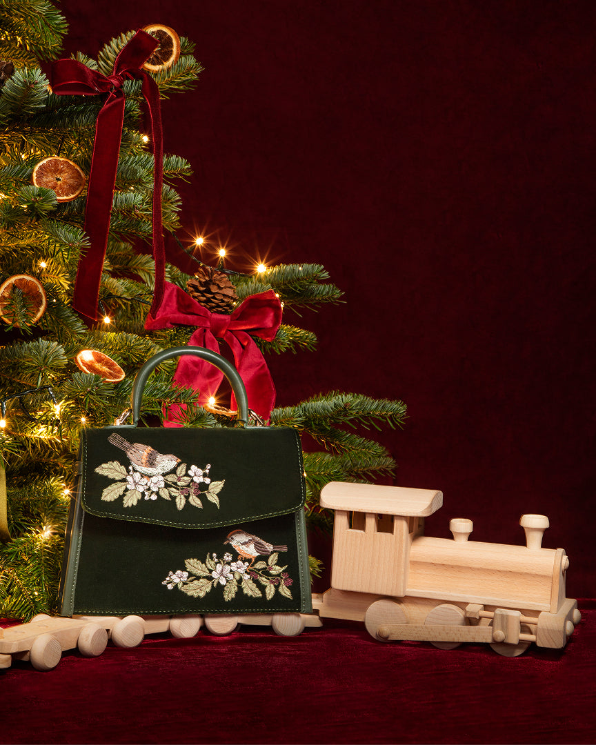 A dark green embroidered handbag with bird and floral motifs, styled on a red velvet surface next to a wooden toy train and a decorated Christmas tree against a dark red background.