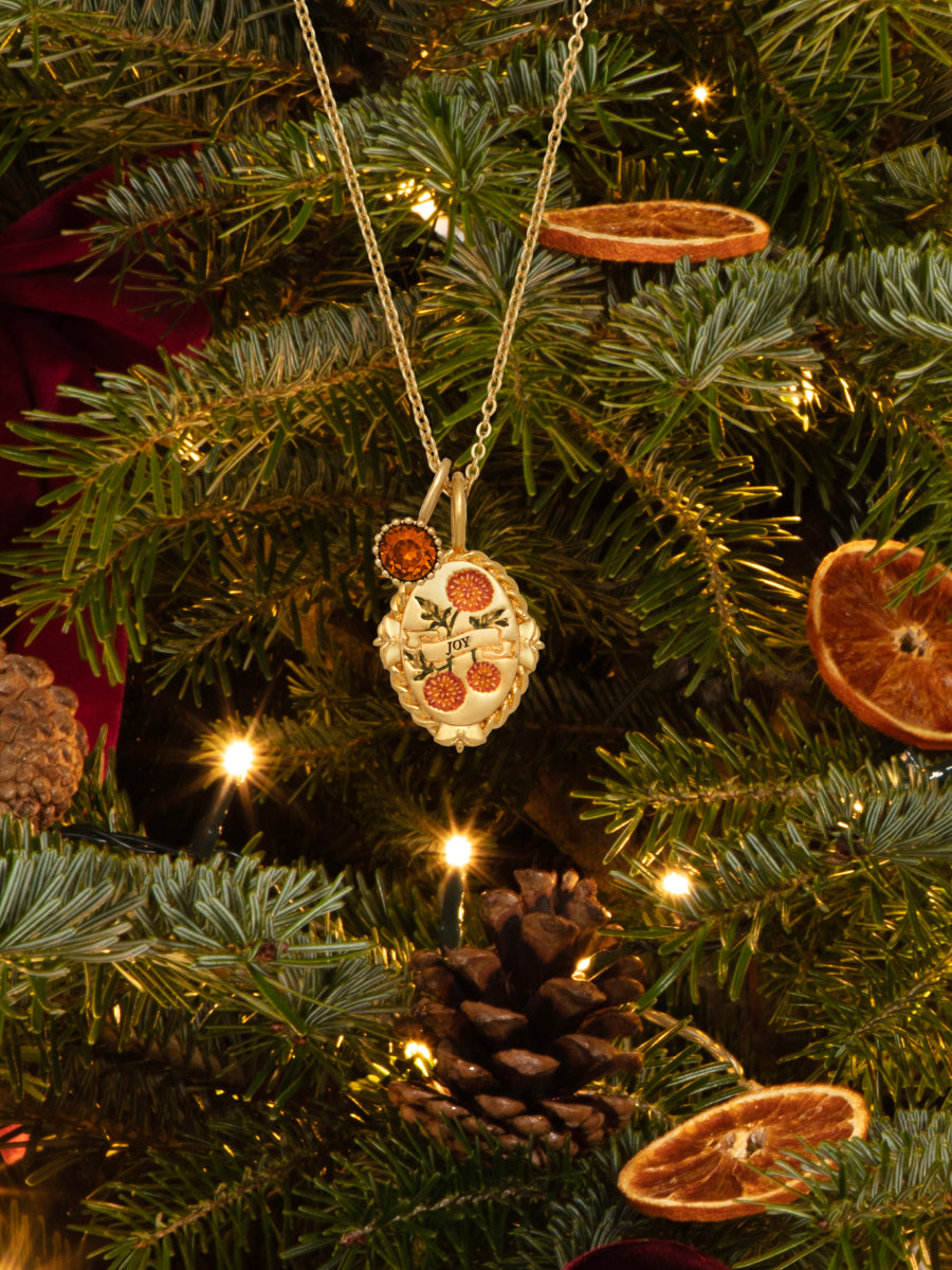 A gold-toned birthstone pendant necklace with a floral design and small orange accents hangs from a Christmas tree decorated with warm lights, pinecones, and dried orange slices.