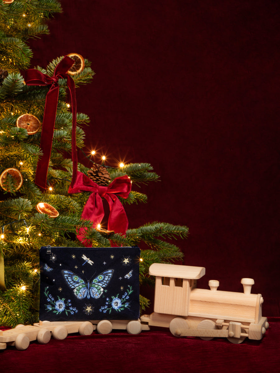 A Christmas-themed scene with a decorated evergreen tree on the left, a red velvet background, a wooden toy train on the right, and a dark gift box with a blue butterfly pouch and floral design in the foreground.