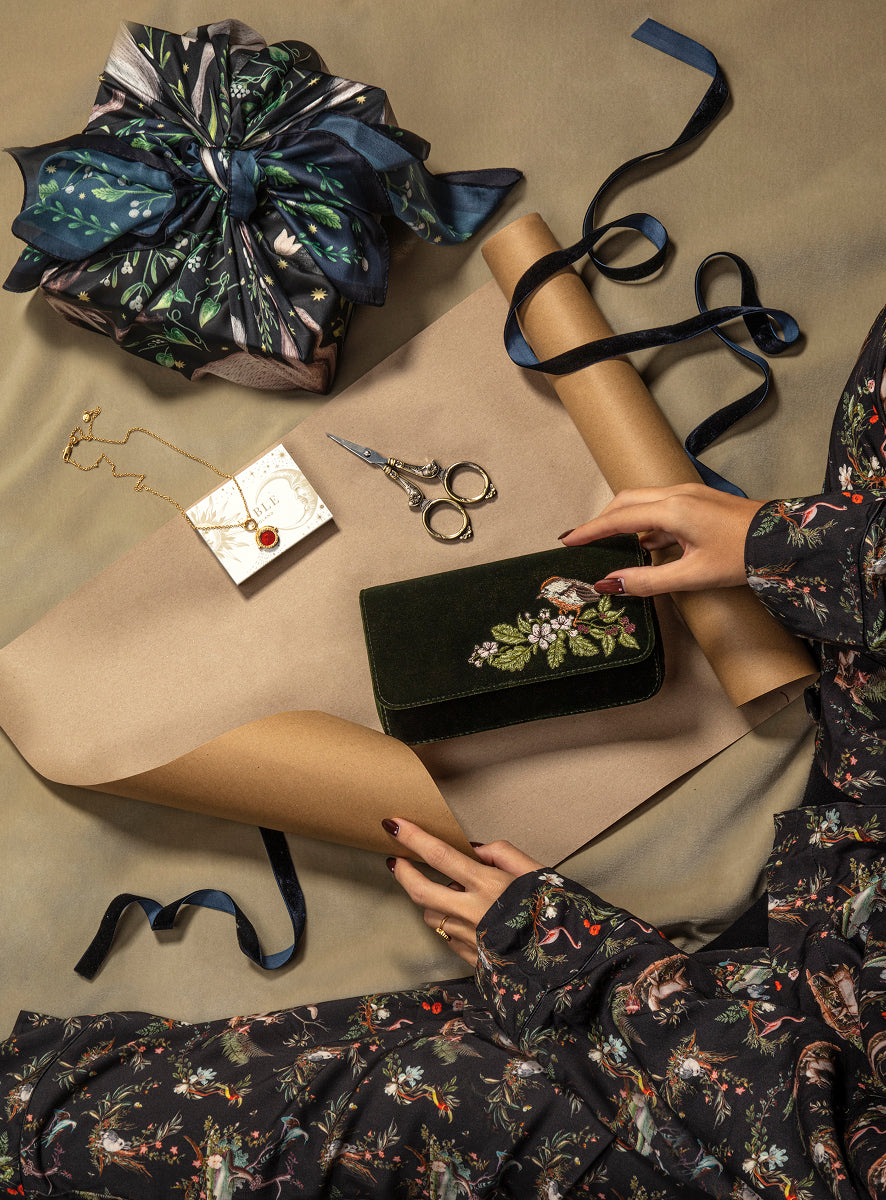 A dark green velvet clutch bag with a floral embroidered design is held by a person wearing a matching dark floral robe, surrounded by wrapping paper, scissors, and a wrapped gift box on a beige surface.