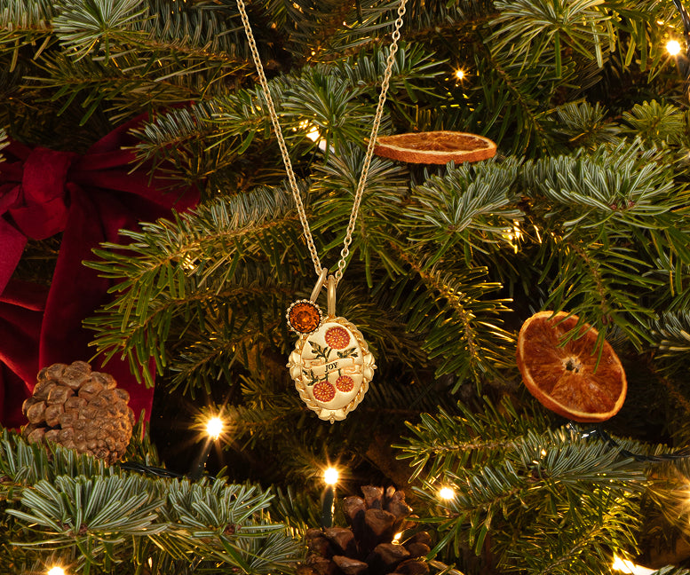 A gold-toned pendant necklace with a floral design hangs from a Christmas tree decorated with green pine branches, warm white string lights, dried orange slices, pinecones, and a red ribbon.