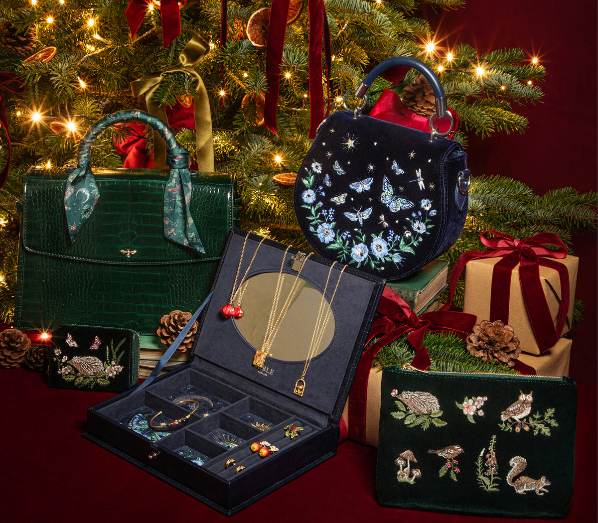 A curated holiday gift set featuring dark green and navy embroidered accessories, including a structured handbag, a round handbag with butterfly embroidery, a jewelry box with necklaces and bracelets, a small pouch with animal embroidery, and wrapped presents, arranged in front of a decorated Christmas tree.
