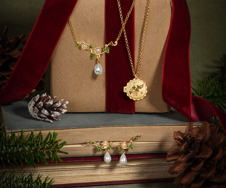 Gold-toned jewelry set displayed on a kraft gift box with red velvet ribbon, including a necklace with a teardrop pearl and a matching locket pendant, plus a matching necklace and pendant laid on books below. Pinecones, evergreen branches, and a dark green background are visible.