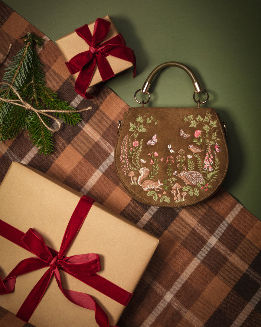 A brown leather handbag with detailed embroidered woodland animals and flowers, styled on a plaid blanket with wrapped gift boxes and evergreen branches.