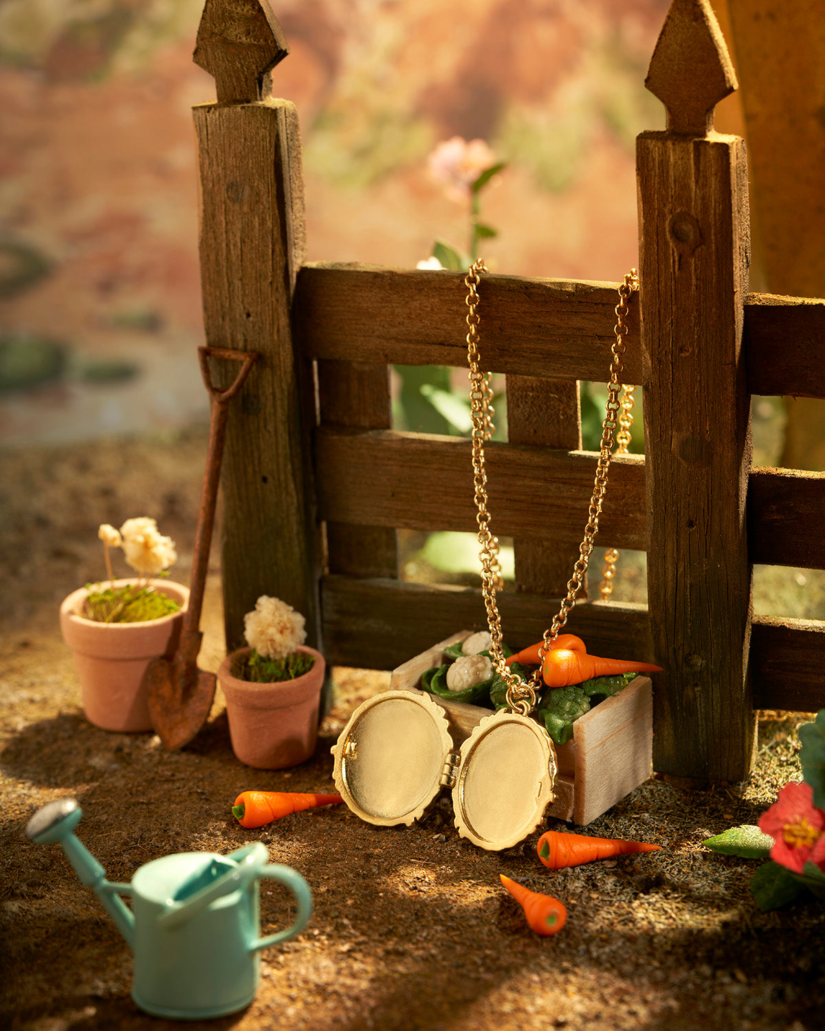 Miniature garden scene with potted plants, carrots, a watering can, and two gold necklaces on a wooden fence with gold locket open.