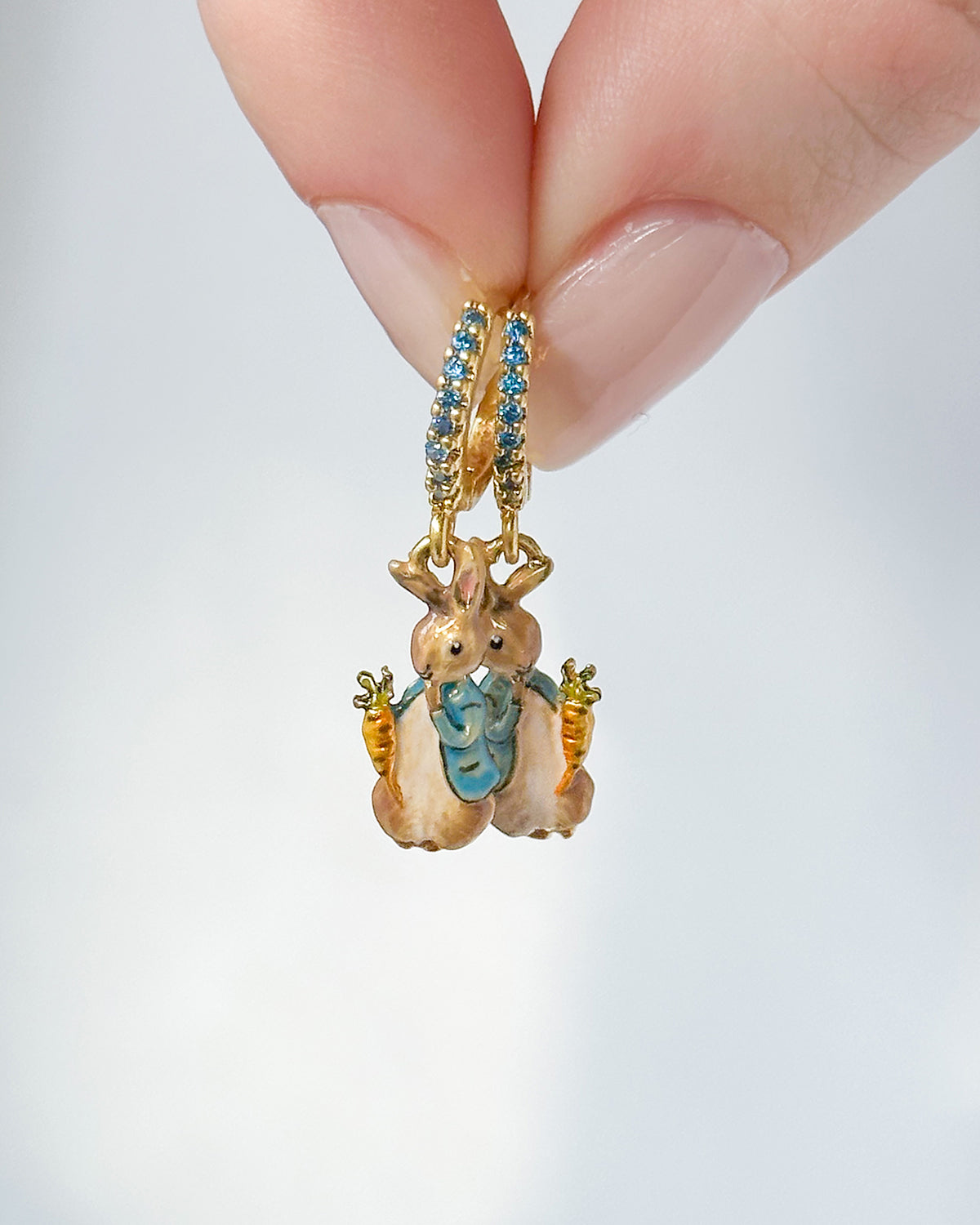 Gold Peter Rabbit charm huggie earrings with blue accents held between fingers on a light gray background