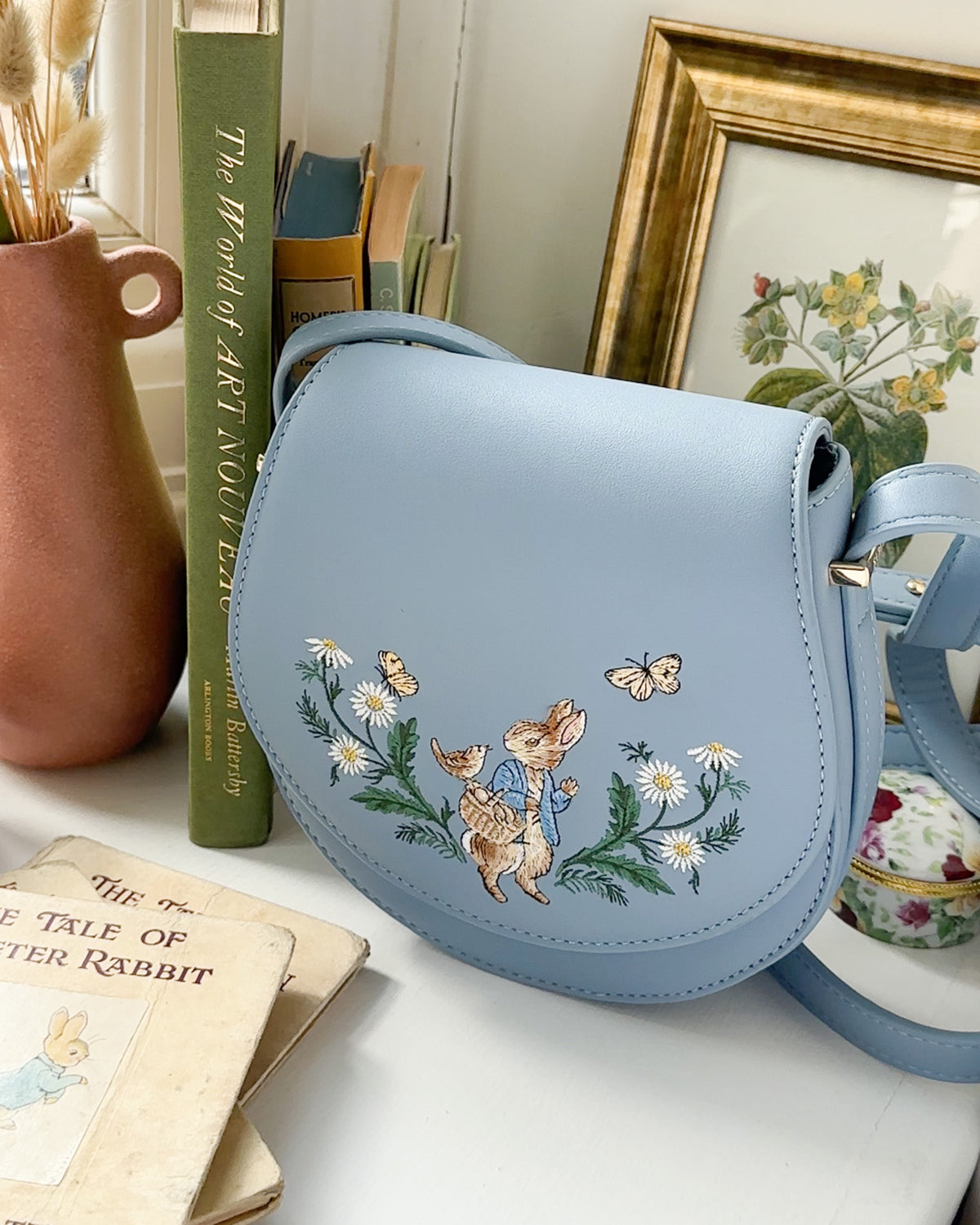Light blue saddle bag with floral embroidery on a surface with books and a teacup in the background