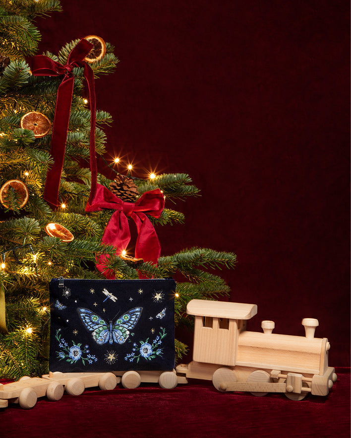 A dark Christmas-themed gift box with a blue butterfly and floral design sits on a red surface next to a wooden toy train, with a decorated Christmas tree in the background.