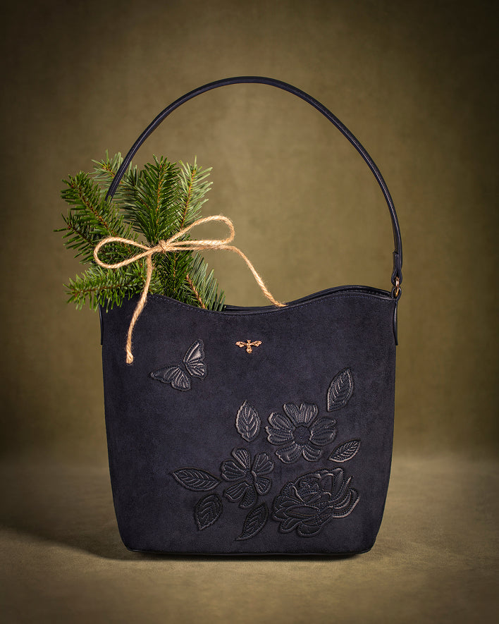 A dark navy blue leather shoulder bag with embossed floral and butterfly details, decorated with a small gold bee emblem and a natural jute twine bow with green pine branches, set against a warm brown textured background.