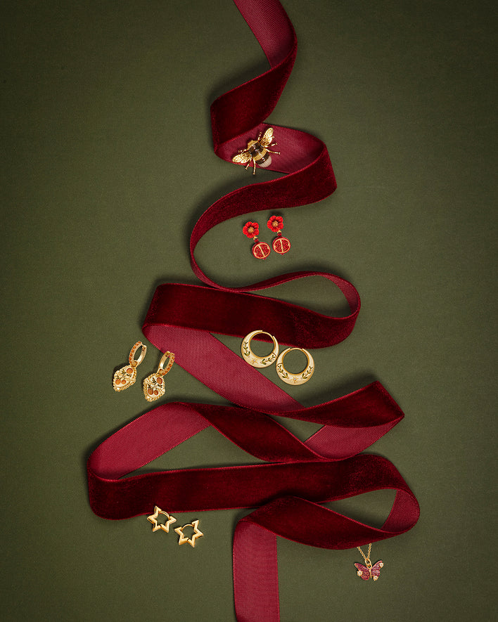 A collection of gold-toned jewelry pieces, including earrings, rings, and a butterfly pendant, arranged on a dark red velvet ribbon that is shaped like a Christmas tree against a dark olive green background.