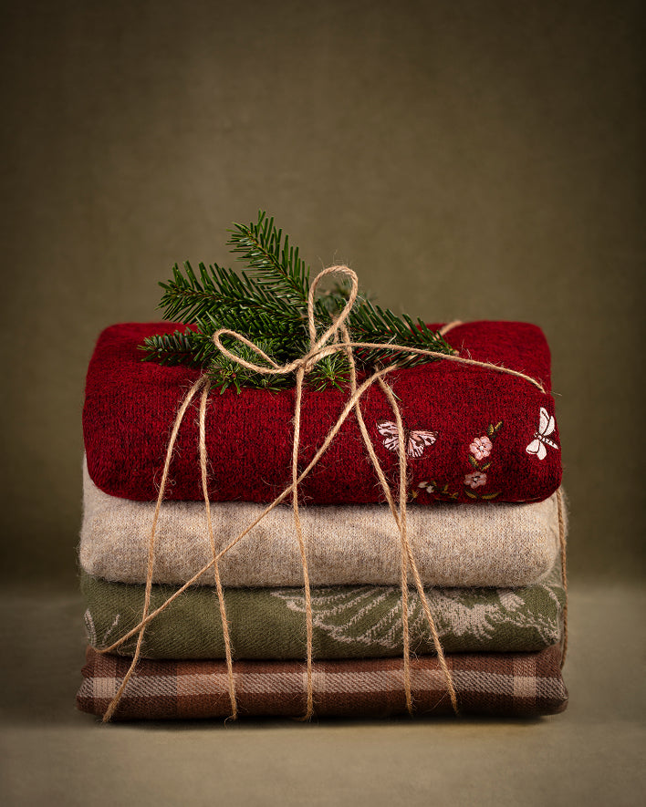 A stack of four folded knit blankets in red, beige, olive green, and brown plaid, wrapped in twine with a green pine sprig on top, set against a dark olive background.