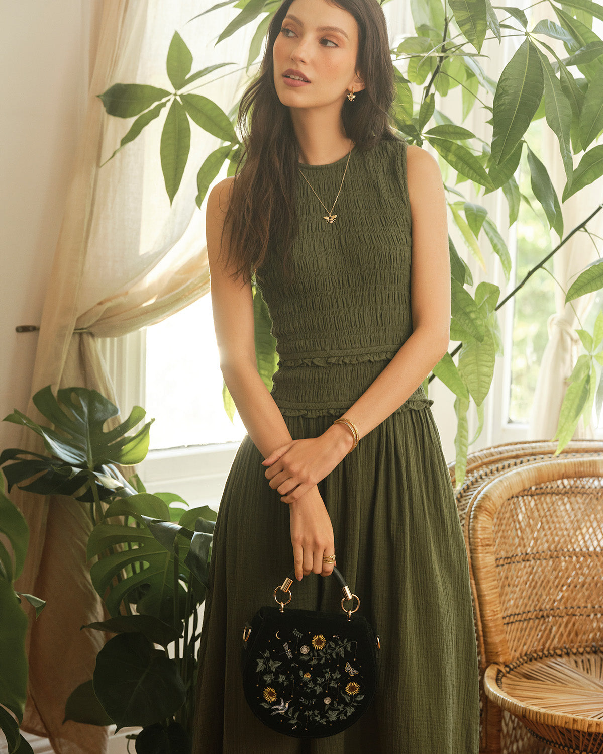 Woman in a green dress holding a dark green handbag with floral details, standing in a room with plants.