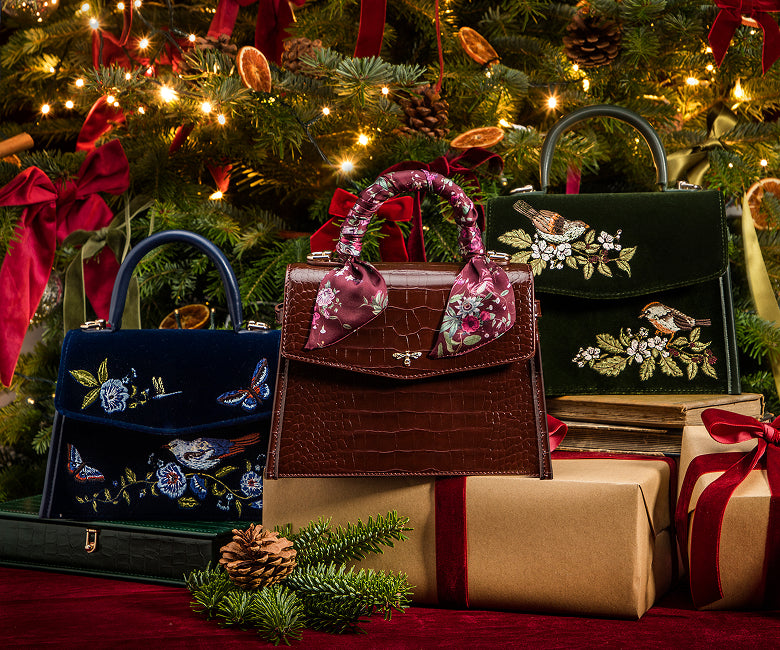 Three decorative handbags arranged in front of a lit Christmas tree with wrapped presents. The central bag is dark brown crocodile-embossed leather with pink floral handles and a dragonfly clasp; the left bag is navy blue velvet with embroidered birds and butterflies; the right bag is dark green velvet with floral embroidery.