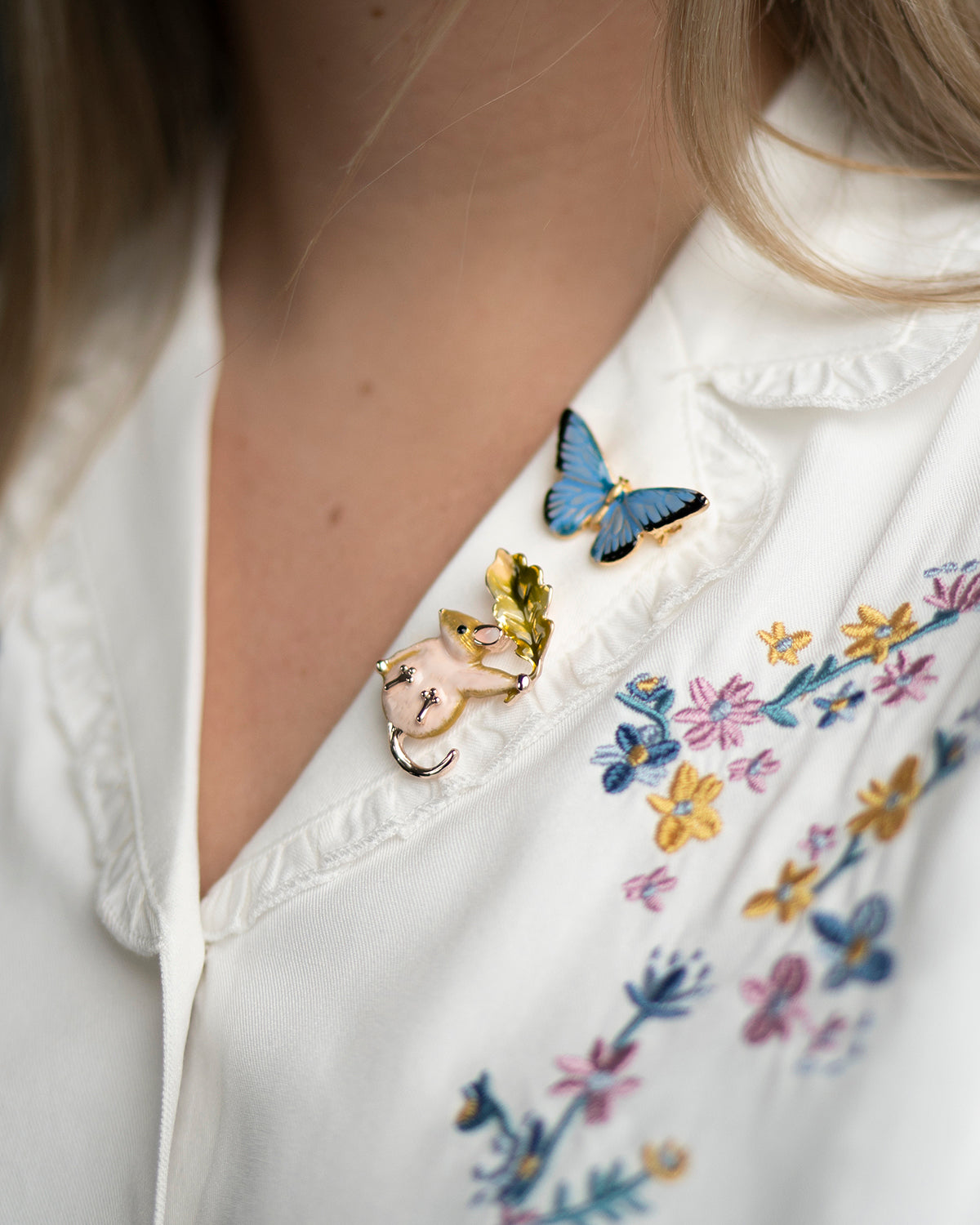 Close-up of a white blouse with floral embroidery and butterfly brooch and dormouse brooch.