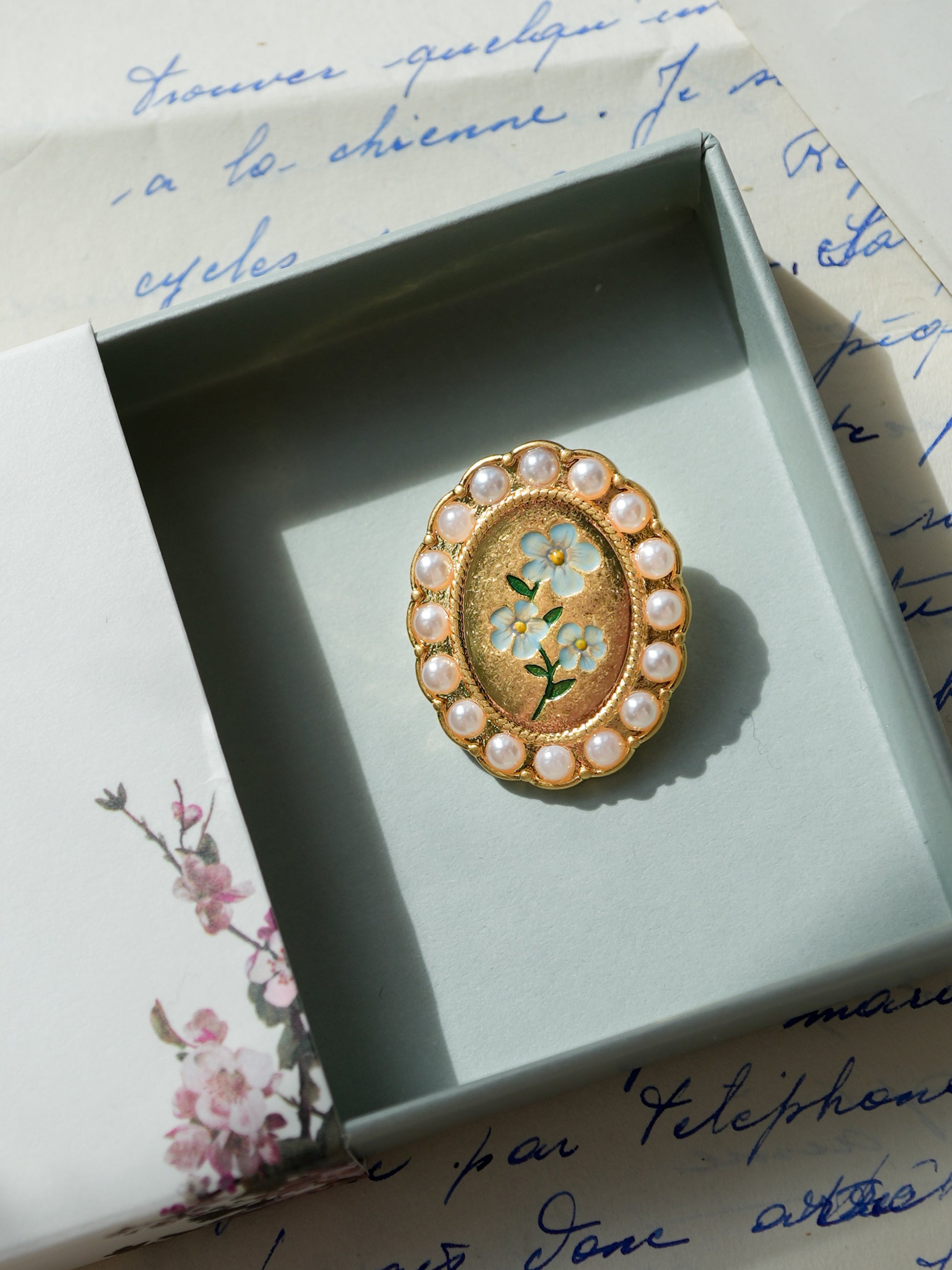Gold brooch with pearl accents in a decorative box on a handwritten letter background