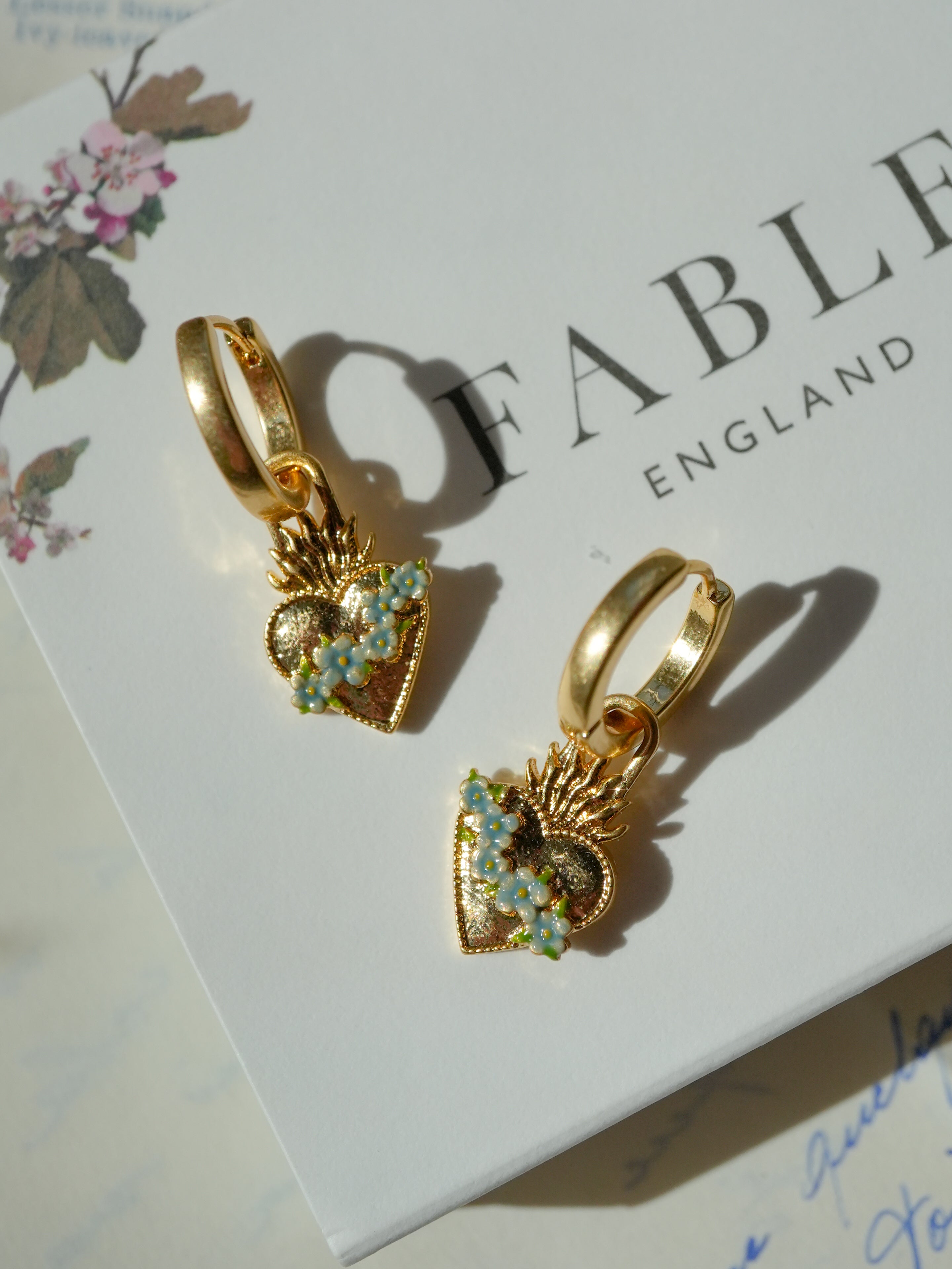 Gold hoop earrings with heart and floral charms on a Fable England box.