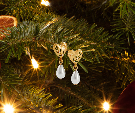 A pair of gold heart-shaped dangle earrings with clear teardrop gemstones, hanging from green Christmas tree branches with warm string lights.
