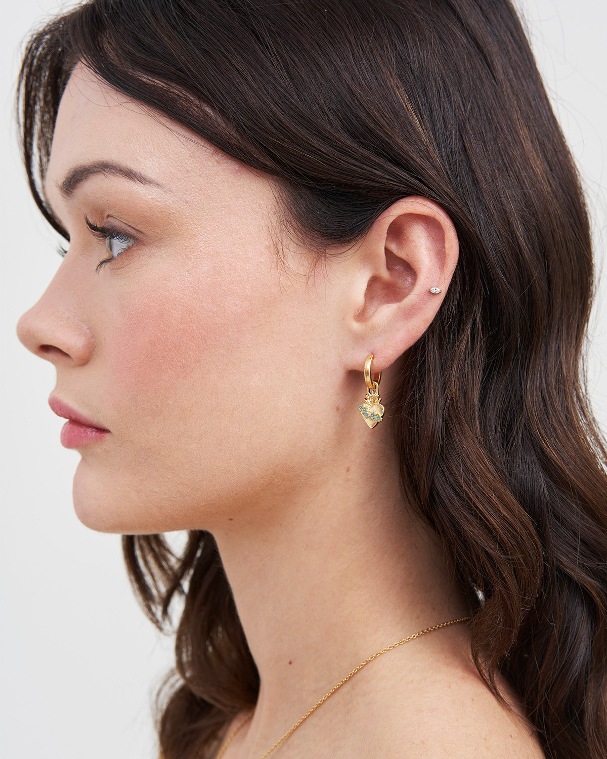 Woman wearing gold sacred heart huggie earrings with a plain background