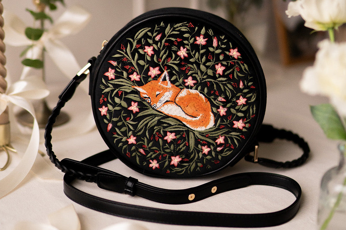 Round embroidered bag with a fox design on a white surface with greenery in the background